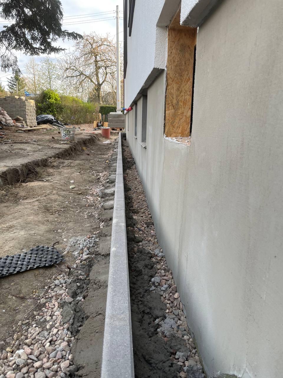 Wąski wykop wzdłuż ściany budynku, wypełniony kamieniami i obrzeżem betonowym. Widoczne przygotowania do drenażu lub izolacji fundamentów. Prace ziemne w toku.
