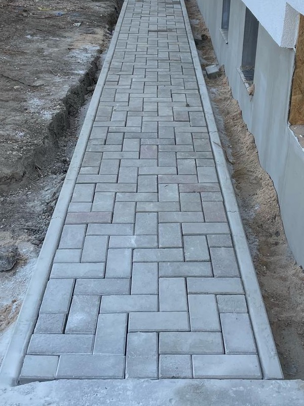 Ścieżka z szarej kostki brukowej ułożona wzdłuż budynku, z widocznym wykopem po lewej stronie. Regularny wzór kostki, betonowe obrzeża, surowe wykończenie elewacji.