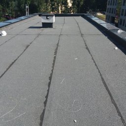 WPIUNIVERSAL - Szary, płaski dach pokryty papą z widocznym kominem i elementami obróbki blacharskiej. Widoczne ślady po naprawach i biały kask. Budynek w tle.