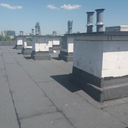 WPIUNIVERSAL - Wykończony dach płaski z kominami wentylacyjnymi z betonu komórkowego, zabezpieczonymi hydroizolacją. W tle panorama miasta w słoneczny dzień.