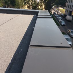 WPIUNIVERSAL - Szczegółowe ujęcie płaskiego dachu z szarą papą i metalowymi obróbkami blacharskimi, widok z góry na krawędź dachu i ulicę z zaparkowanymi autami.