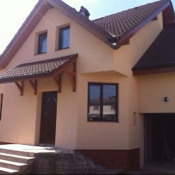 WPIUNIVERSAL - Elewacja domu jednorodzinnego w beżowym kolorze z brązowymi oknami i dachem. Widoczny fragment garażu i schody wejściowe.