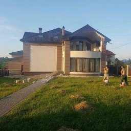 WPIUNIVERSAL - Dwukondygnacyjny dom z jasną elewacją w trakcie malowania. Trzech mężczyzn pracuje na trawniku przed budynkiem, używając wiader z farbą. Widoczne ślady po pracach.