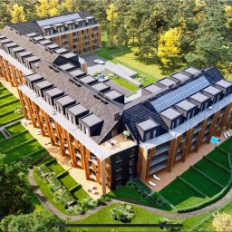 WPIUNIVERSAL - Wizualizacja z lotu ptaka nowoczesnego apartamentowca z panelami słonecznymi na dachu, otoczonego zielenią i drzewami. Widoczne balkony i tarasy.
