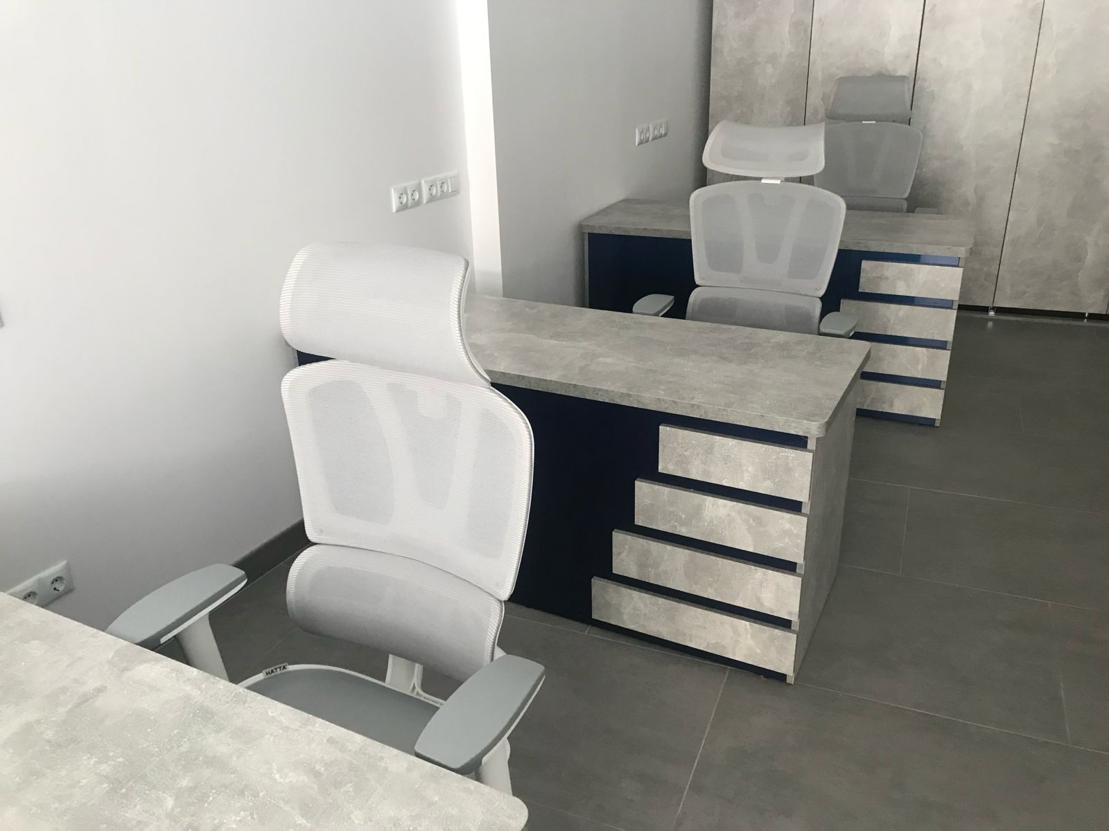 Nowoczesne biuro z szarymi biurkami z szufladami i białymi krzesłami biurowymi z siatkowym oparciem. Minimalistyczny design, neutralna kolorystyka, widoczne gniazdka elektryczne.