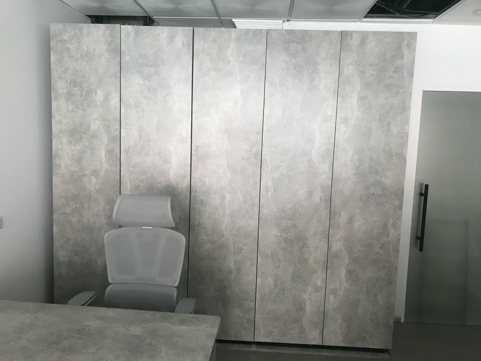 Szafa na wymiar z szarymi frontami imitującymi kamień, z krzesłem biurowym i fragmentem biurka w minimalistycznym wnętrzu biura. Widoczne oświetlenie sufitowe.