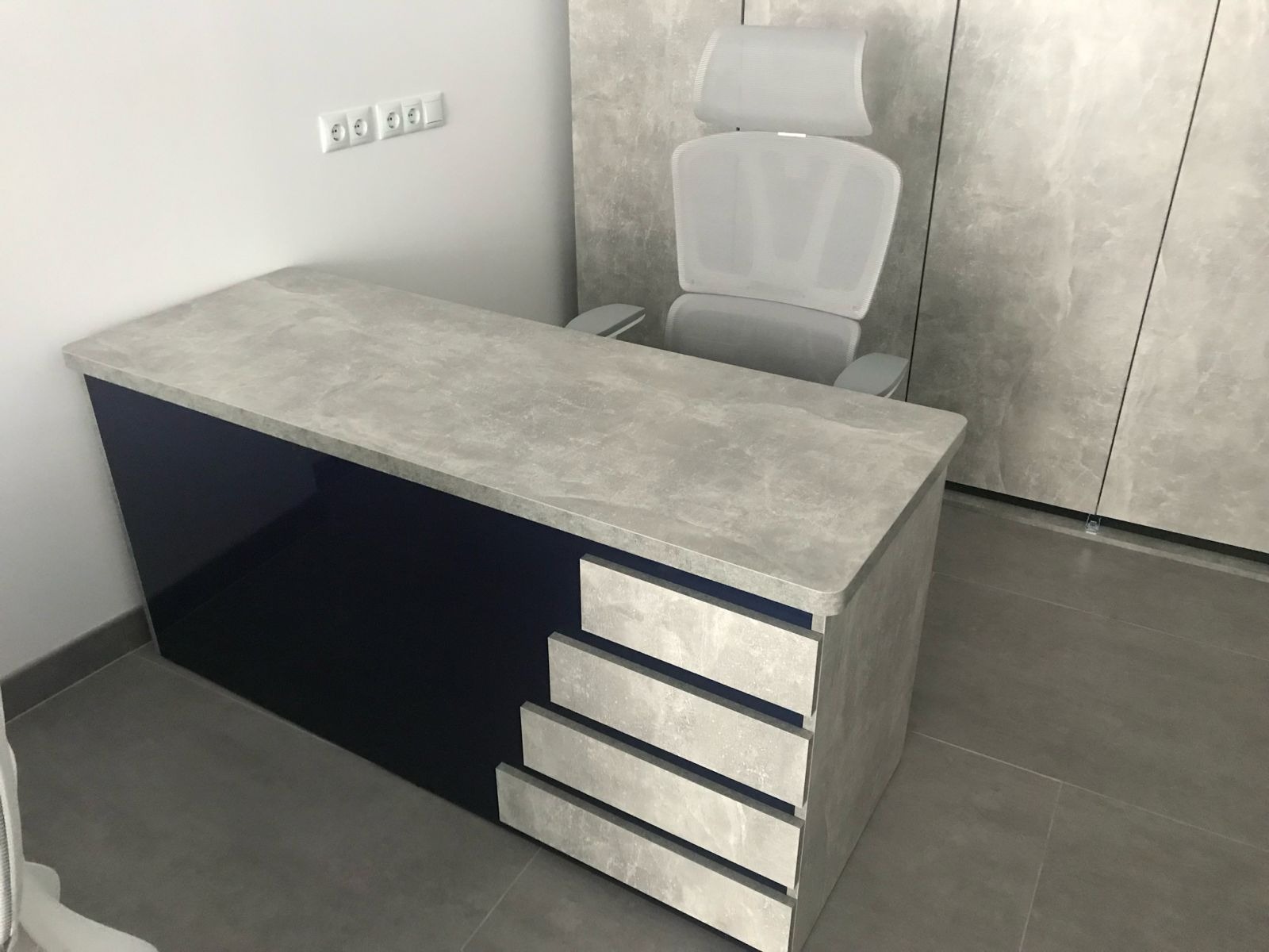 Biurko z szufladami i krzesłem w nowoczesnym biurze. Blat biurka imituje beton, fronty szuflad i bok biurka w kolorze szarym, a bok biurka w kolorze granatowym.