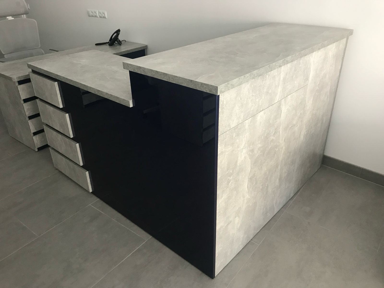 Nowoczesna lada recepcyjna z czarnymi frontami i blatem imitującym beton, uzupełniona o szuflady w tym samym stylu. Minimalistyczny design, widok z perspektywy.