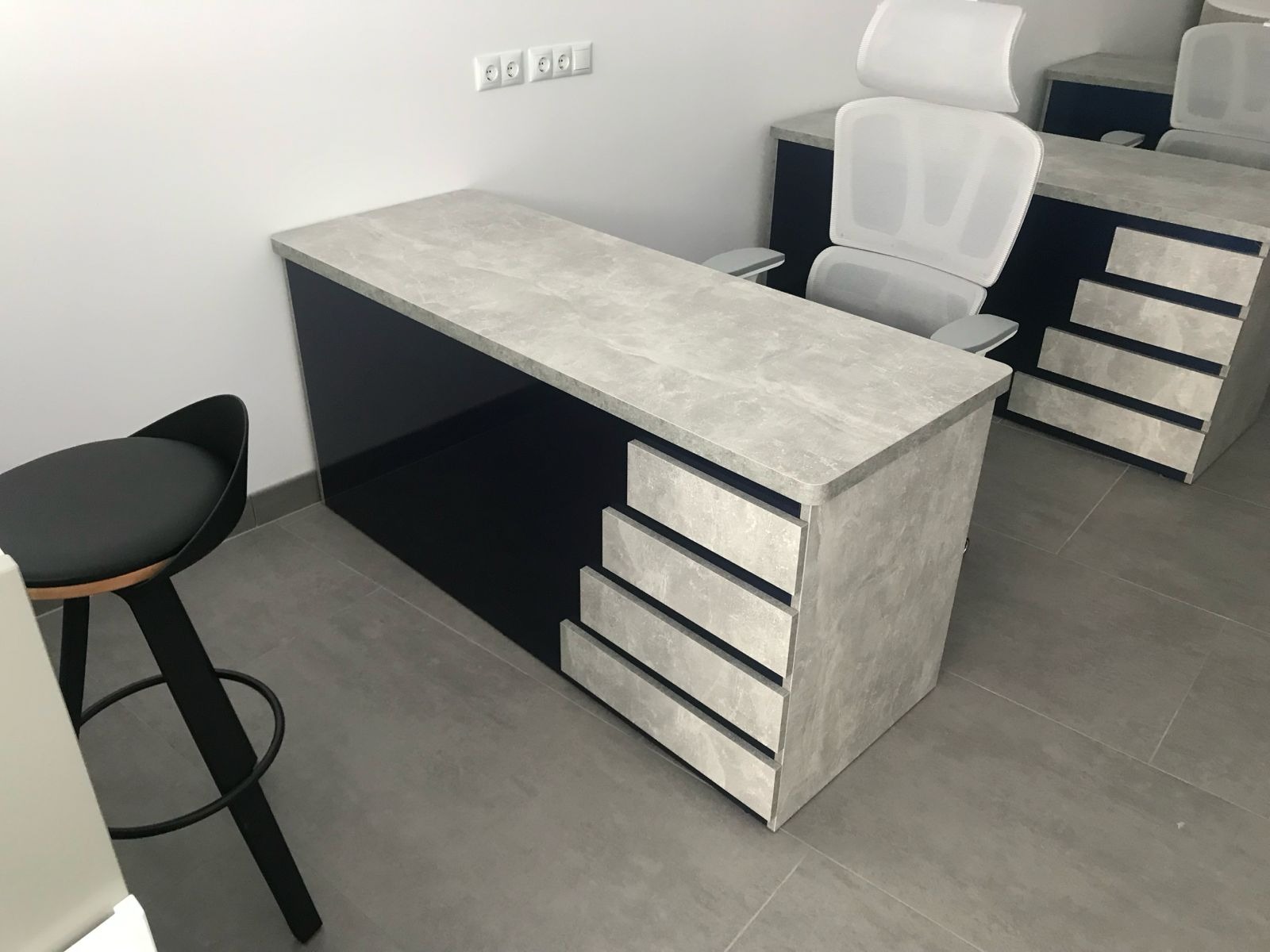 Nowoczesne biurko z szufladami w odcieniach szarości i granatu, z krzesłem barowym i biurowym w tle, na tle białej ściany z gniazdkami.