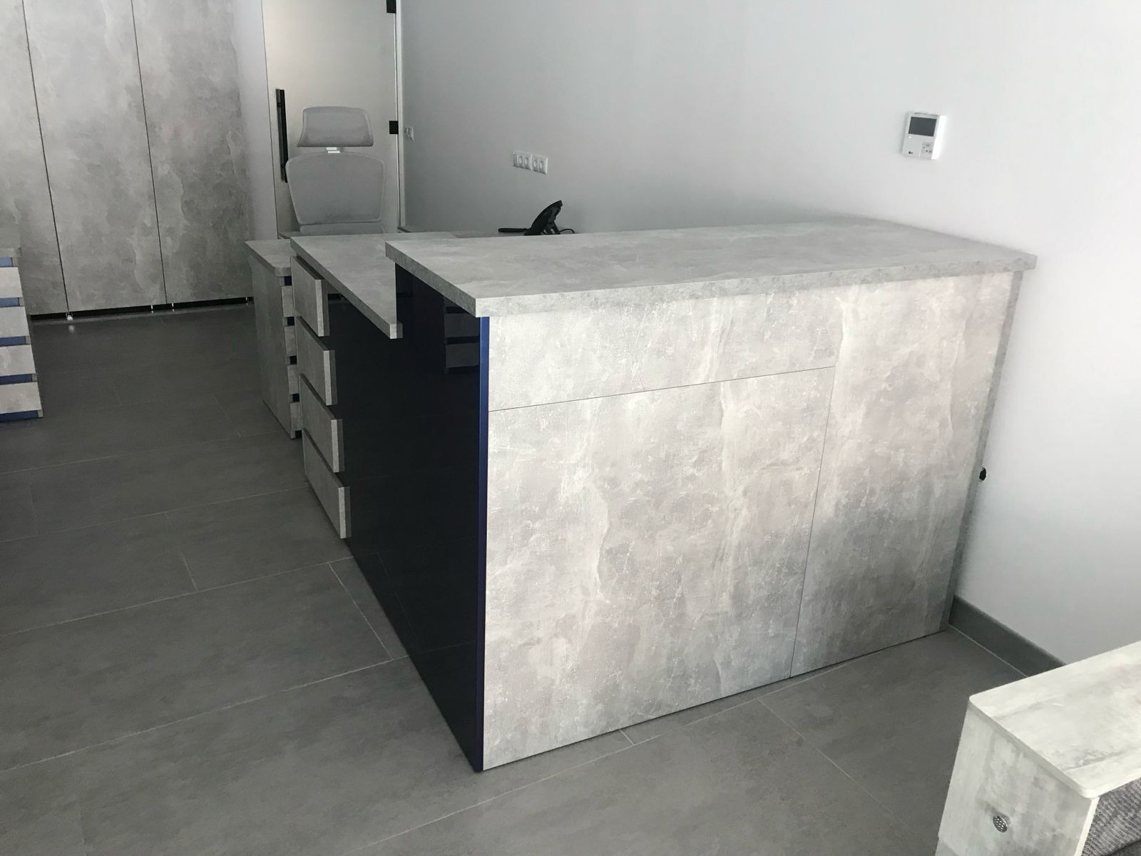 Nowoczesna, szara lada recepcyjna z ciemnymi elementami, wykonana na wymiar. Widoczne szuflady i wykończenie imitujące kamień. Minimalistyczny design.