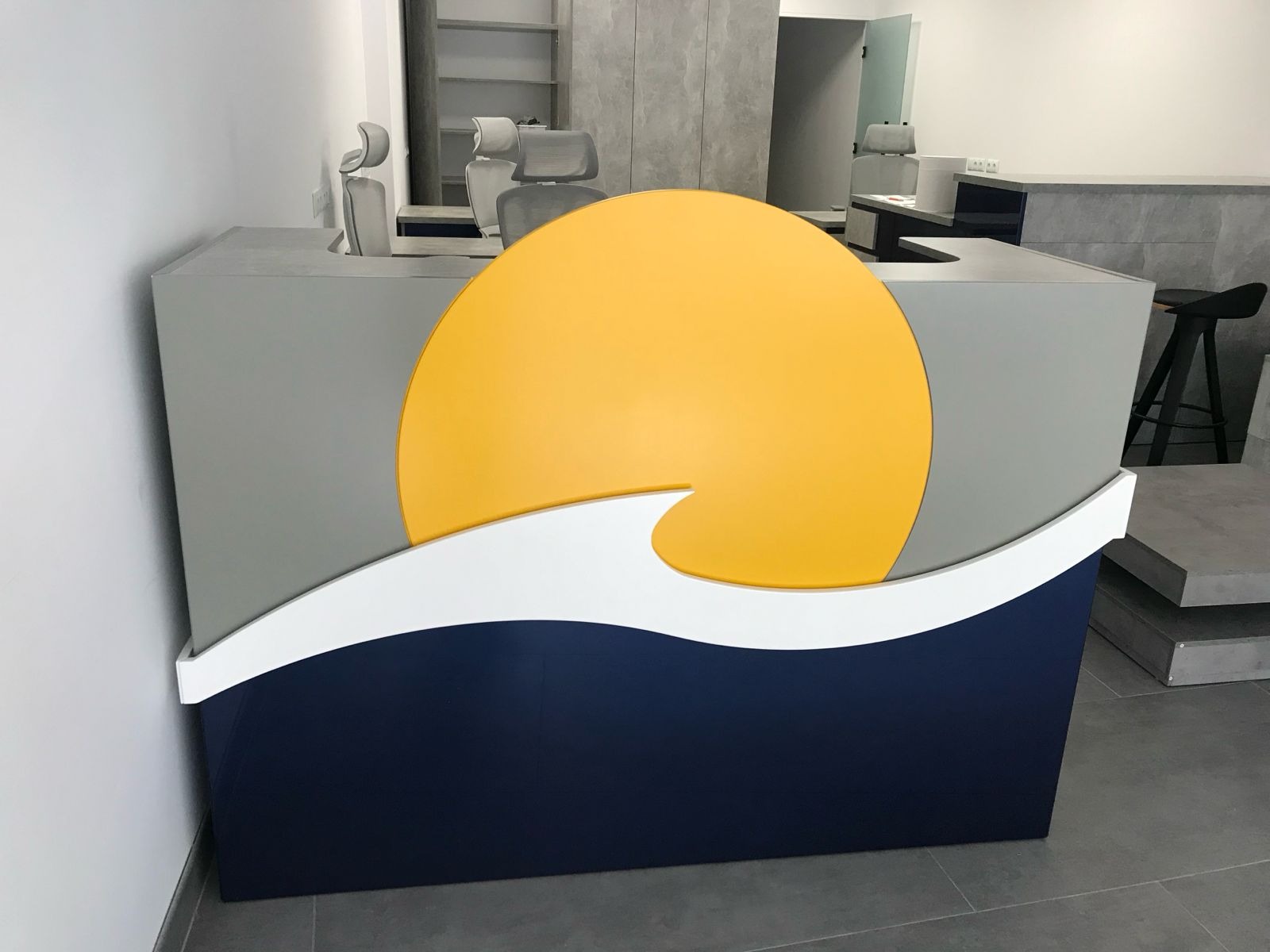 Recepcja z motywem słońca i fali w kolorach żółtym, białym i granatowym. Szare ściany i podłoga. W tle krzesła i biurka.