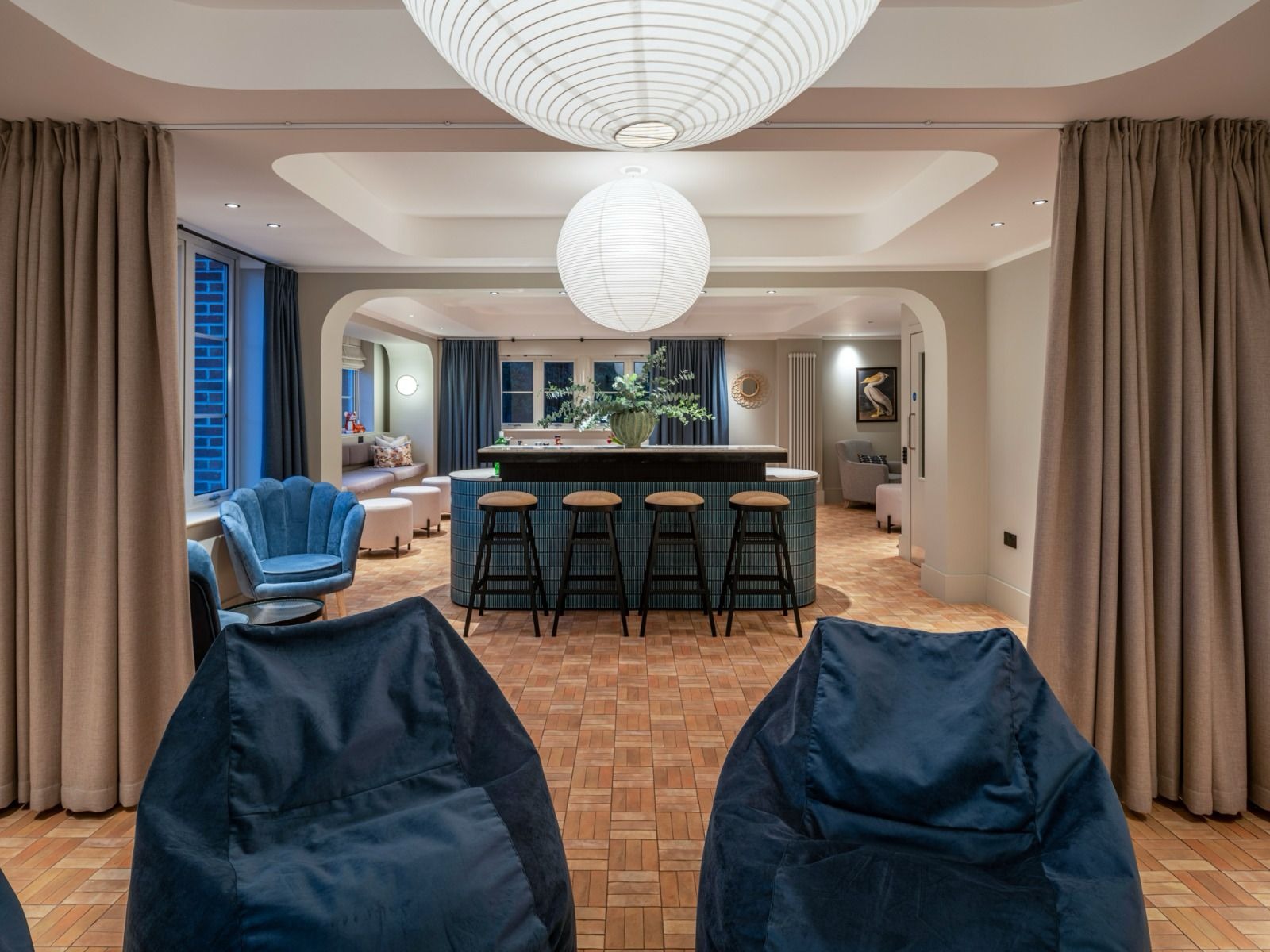 Stylowe wnętrze z barem, krzesłami barowymi i pufami. Uwagę przykuwają lampy w kształcie kul oraz parkiet ułożony w kwadraty. Widoczne zasłony i dekoracje.