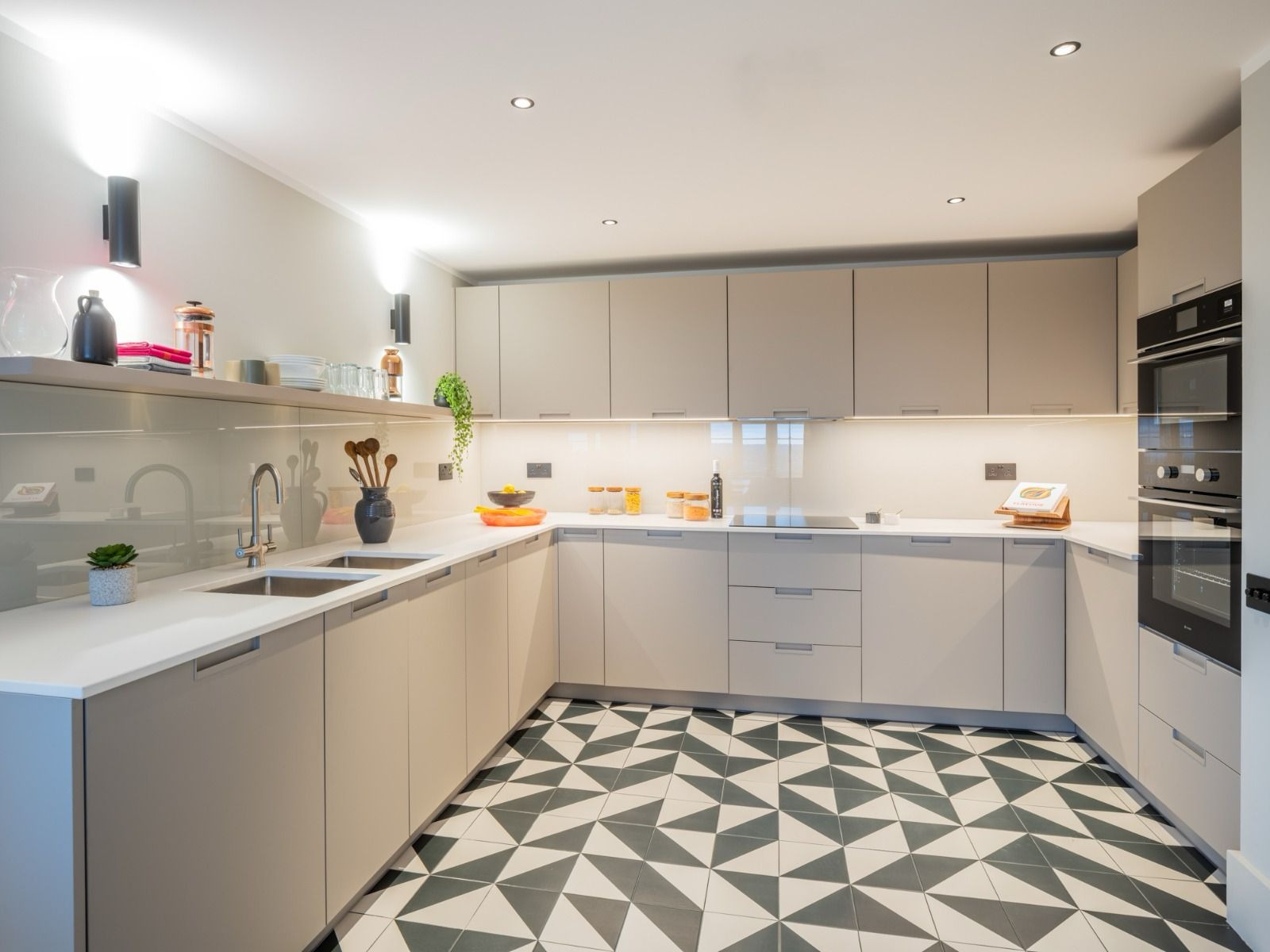 Jasna kuchnia w kształcie litery U z geometryczną podłogą. Minimalistyczne szafki, oświetlenie LED pod szafkami i czarne lampy ścienne. Nowoczesny design z elementami dekoracyjnymi.