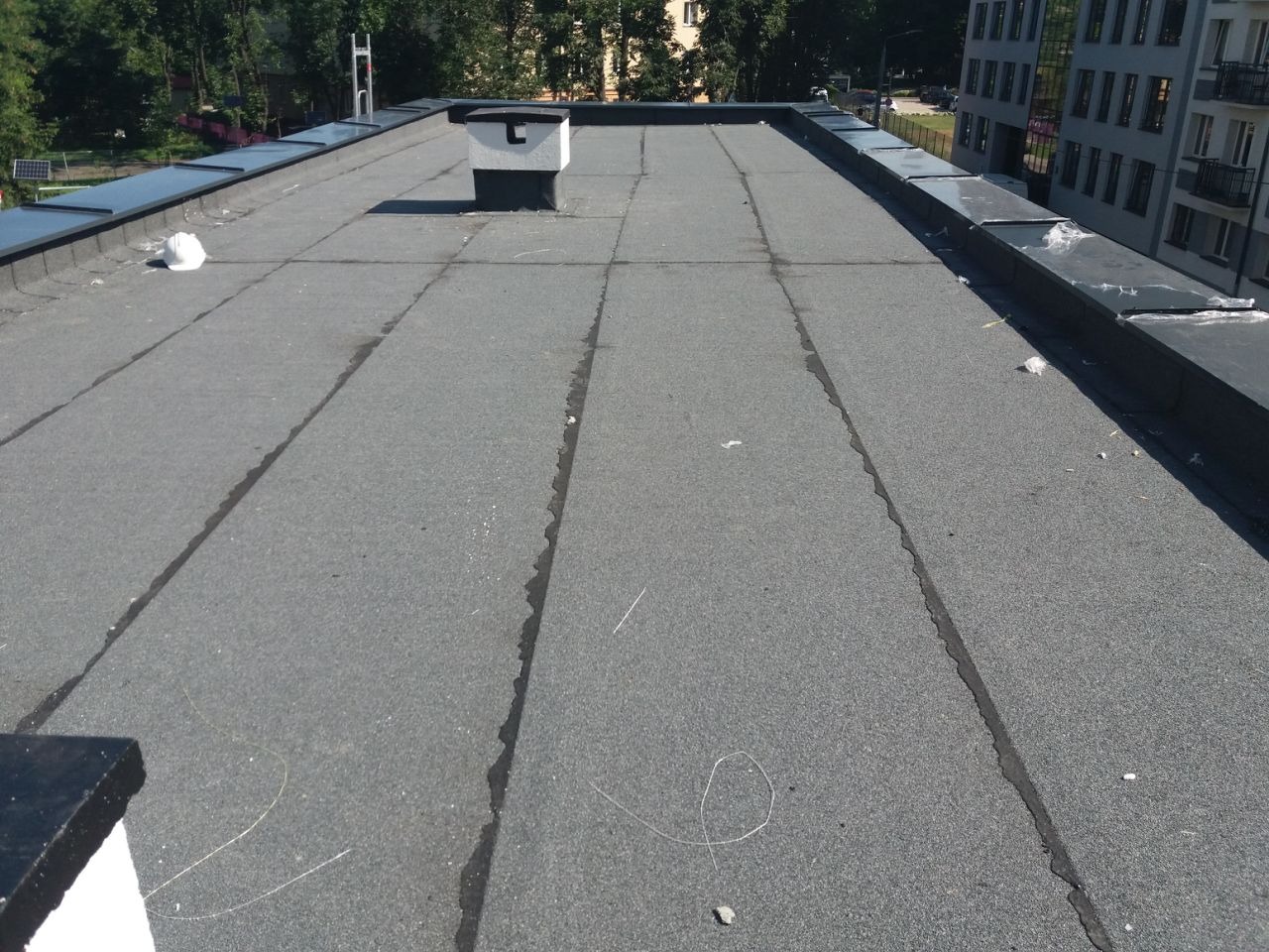 Szary, płaski dach pokryty papą z widocznym kominem i elementami obróbki blacharskiej. Widoczne ślady po naprawach i biały kask. Budynek w tle.