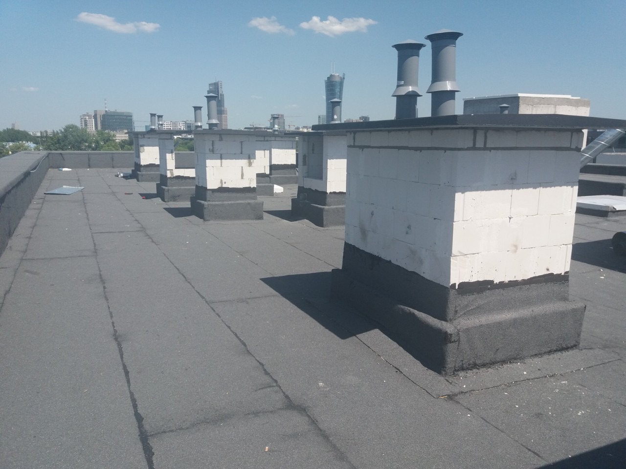 Wykończony dach płaski z kominami wentylacyjnymi z betonu komórkowego, zabezpieczonymi hydroizolacją. W tle panorama miasta w słoneczny dzień.