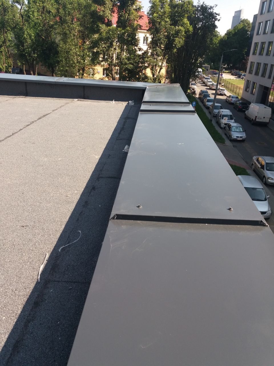 Szczegółowe ujęcie płaskiego dachu z szarą papą i metalowymi obróbkami blacharskimi, widok z góry na krawędź dachu i ulicę z zaparkowanymi autami.