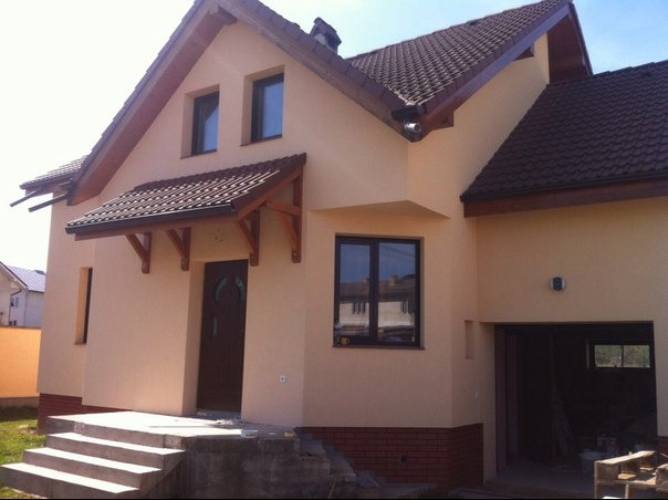 Elewacja domu jednorodzinnego w beżowym kolorze z brązowymi oknami i dachem. Widoczny fragment garażu i schody wejściowe.