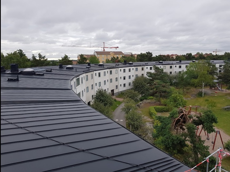 Czarny, metalowy dach wielorodzinnego budynku z widokiem na park i plac zabaw w tle. Ujęcie z góry, w tle żuraw budowlany.