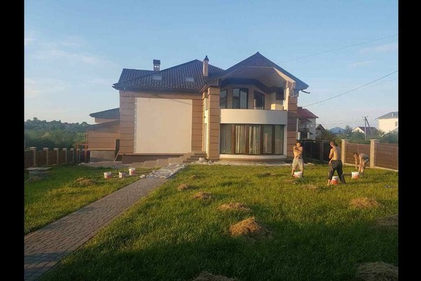 Dwukondygnacyjny dom z jasną elewacją w trakcie malowania. Trzech mężczyzn pracuje na trawniku przed budynkiem, używając wiader z farbą. Widoczne ślady po pracach.