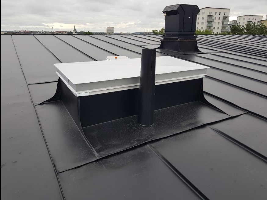 Czarny, blaszany dach z kominem i lukarną w kolorze białym. W tle zabudowa miejska pod pochmurnym niebem. Widok z góry na nowo położone pokrycie dachowe.