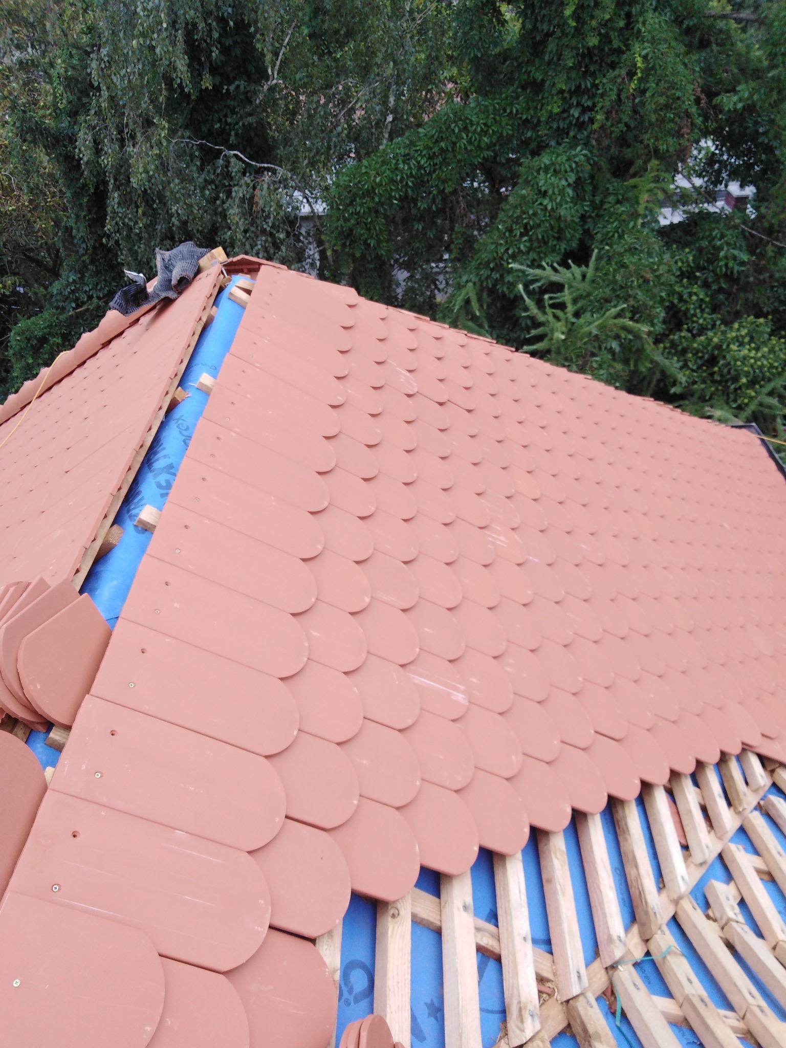 Układanie dachówki karpiówki w łuskę na niebieskiej membranie. Widoczne drewniane łaty i fragment zieleni w tle. Dach w trakcie krycia.