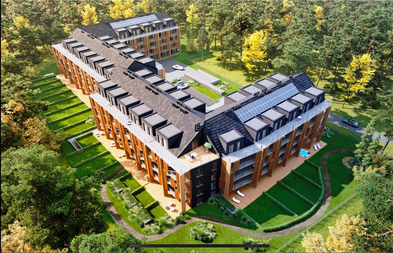 Wizualizacja z lotu ptaka nowoczesnego apartamentowca z panelami słonecznymi na dachu, otoczonego zielenią i drzewami. Widoczne balkony i tarasy.