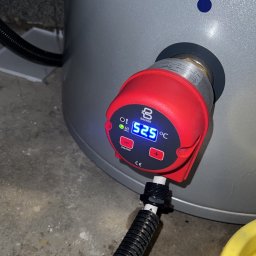 WM POMPY CIEPŁA I KLIMATYZACJA - Szary zbiornik z czerwonym regulatorem temperatury Backer, wyświetlającym 52.5°C. Widoczny fragment instalacji i podłogi. Ujęcie z bliska, skupione na detalu.