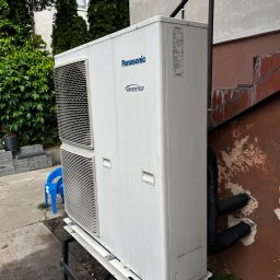 WM POMPY CIEPŁA I KLIMATYZACJA - Zewnętrzna jednostka klimatyzacji Panasonic Inverter, zamontowana na metalowej konstrukcji przy ścianie budynku. Widoczne rury w izolacji wchodzące w ścianę.