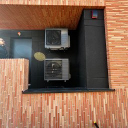 WM POMPY CIEPŁA I KLIMATYZACJA - Dwa klimatyzatory zamontowane na elewacji z cegły klinkierowej, czarne panele pod urządzeniami. Widoczny fragment osoby w okularach po lewej stronie.