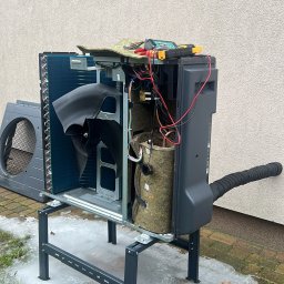 WM POMPY CIEPŁA I KLIMATYZACJA - Otwarta obudowa pompy ciepła na zewnątrz budynku, widoczne elementy wewnętrzne i narzędzia diagnostyczne, otoczenie z resztkami śniegu.