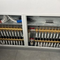 WM POMPY CIEPŁA I KLIMATYZACJA - Rozdzielacz ogrzewania podłogowego w metalowej obudowie, widok z góry. Dwa rzędy rur z numeracją, przewody elektryczne w tle. Instalacja w kotłowni.