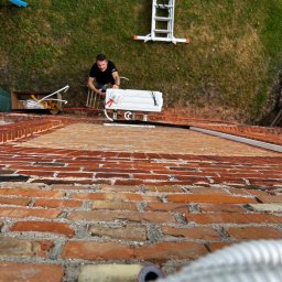 WM POMPY CIEPŁA I KLIMATYZACJA - Montaż klimatyzacji na dachu z cegły. Instalator na drabinie przygotowuje jednostkę zewnętrzną. Widok z góry, z perspektywy dachu, na trawnik i narzędzia.