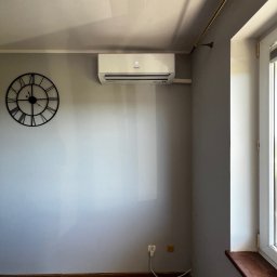 WM POMPY CIEPŁA I KLIMATYZACJA - Biały klimatyzator ścienny w szarym pokoju z drewnianą podłogą, widoczny zegar na ścianie i okno z parapetem. Minimalistyczny design wnętrza.