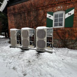 WM POMPY CIEPŁA I KLIMATYZACJA - Trzy jednostki zewnętrzne pompy ciepła, częściowo zasypane śniegiem, ustawione przed ceglanym budynkiem z oknem z zielonymi okiennicami z motywem konia.