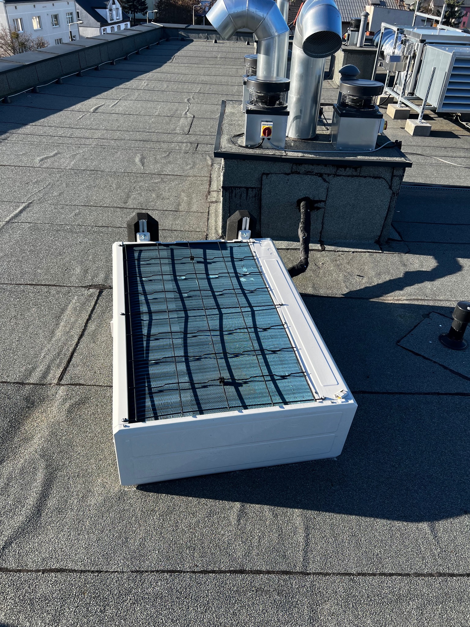 Biała jednostka zewnętrzna klimatyzacji na płaskim dachu z szarej papy, widoczne przewody wentylacyjne i inne urządzenia. Słoneczny dzień.