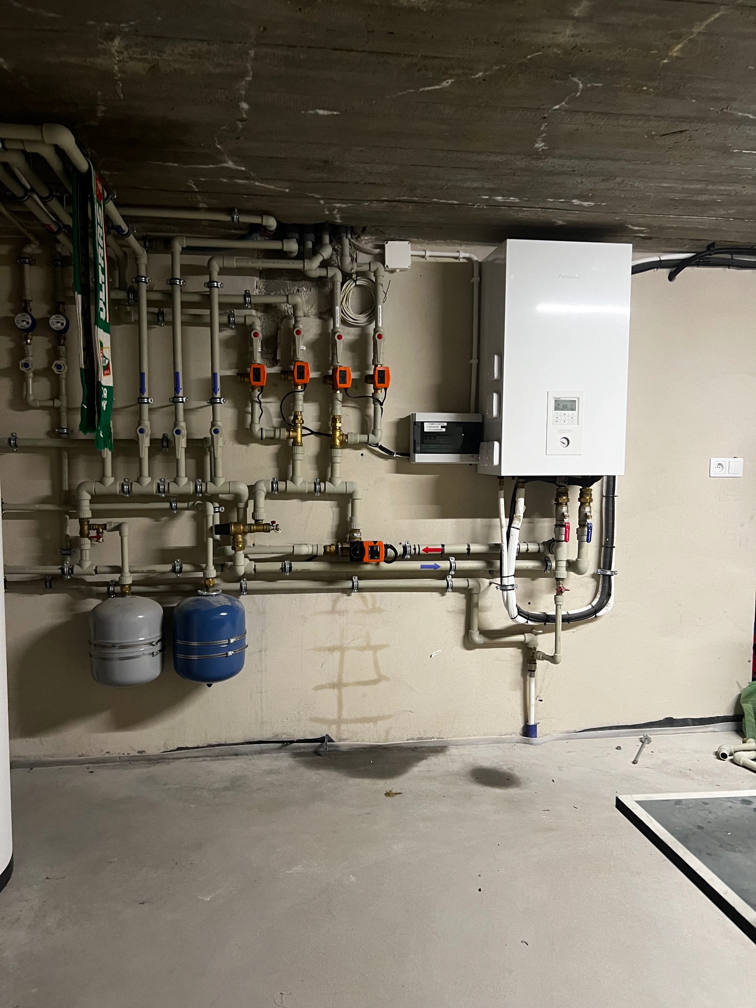 Biały piec grzewczy zamontowany na ścianie w piwnicy, z widoczną instalacją hydrauliczną, rurami i zbiornikami wyrównawczymi. Surowy betonowy sufit i szara podłoga.