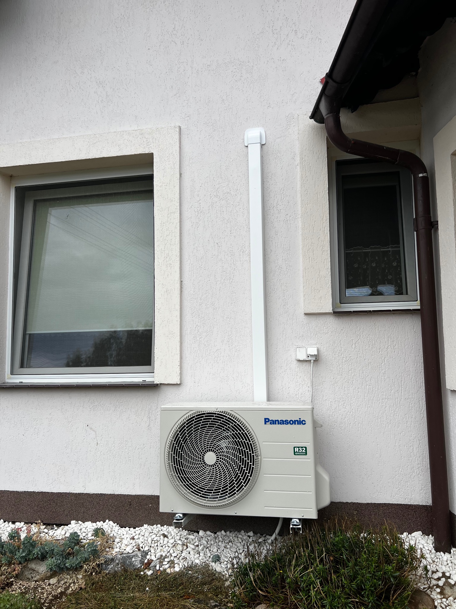 Zewnętrzna jednostka klimatyzacji Panasonic R32 zamontowana na elewacji budynku z oknami, rurą odprowadzającą i kamieniami dekoracyjnymi u podstawy.