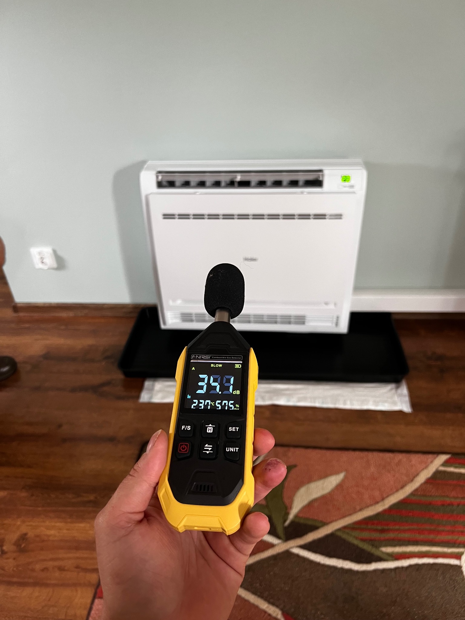 Pomiar głośności klimatyzatora ściennego za pomocą sonometru. Żółty miernik w dłoni pokazuje 39.9 dB. Biały klimatyzator na ścianie w tle.