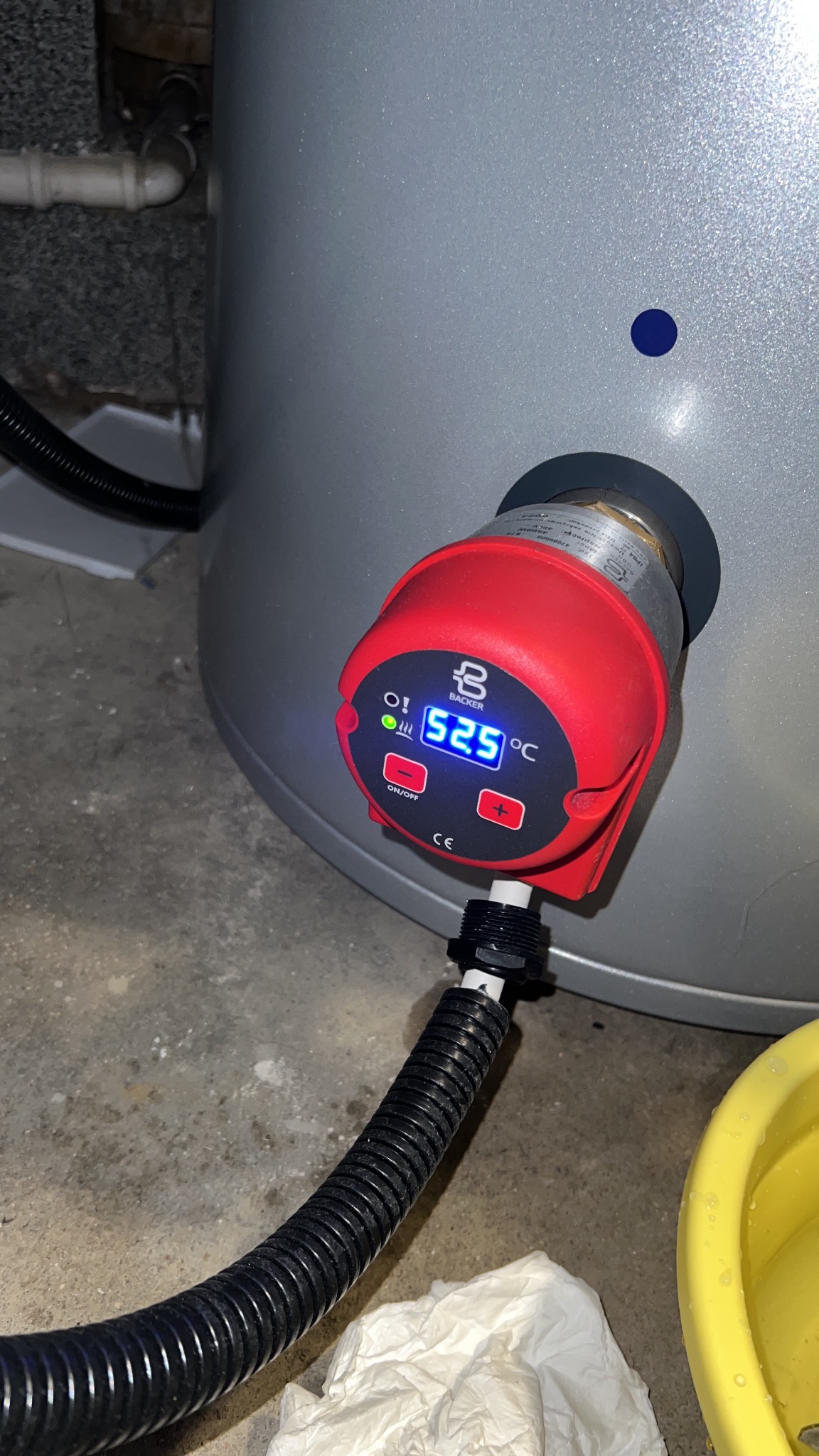 Szary zbiornik z czerwonym regulatorem temperatury Backer, wyświetlającym 52.5°C. Widoczny fragment instalacji i podłogi. Ujęcie z bliska, skupione na detalu.