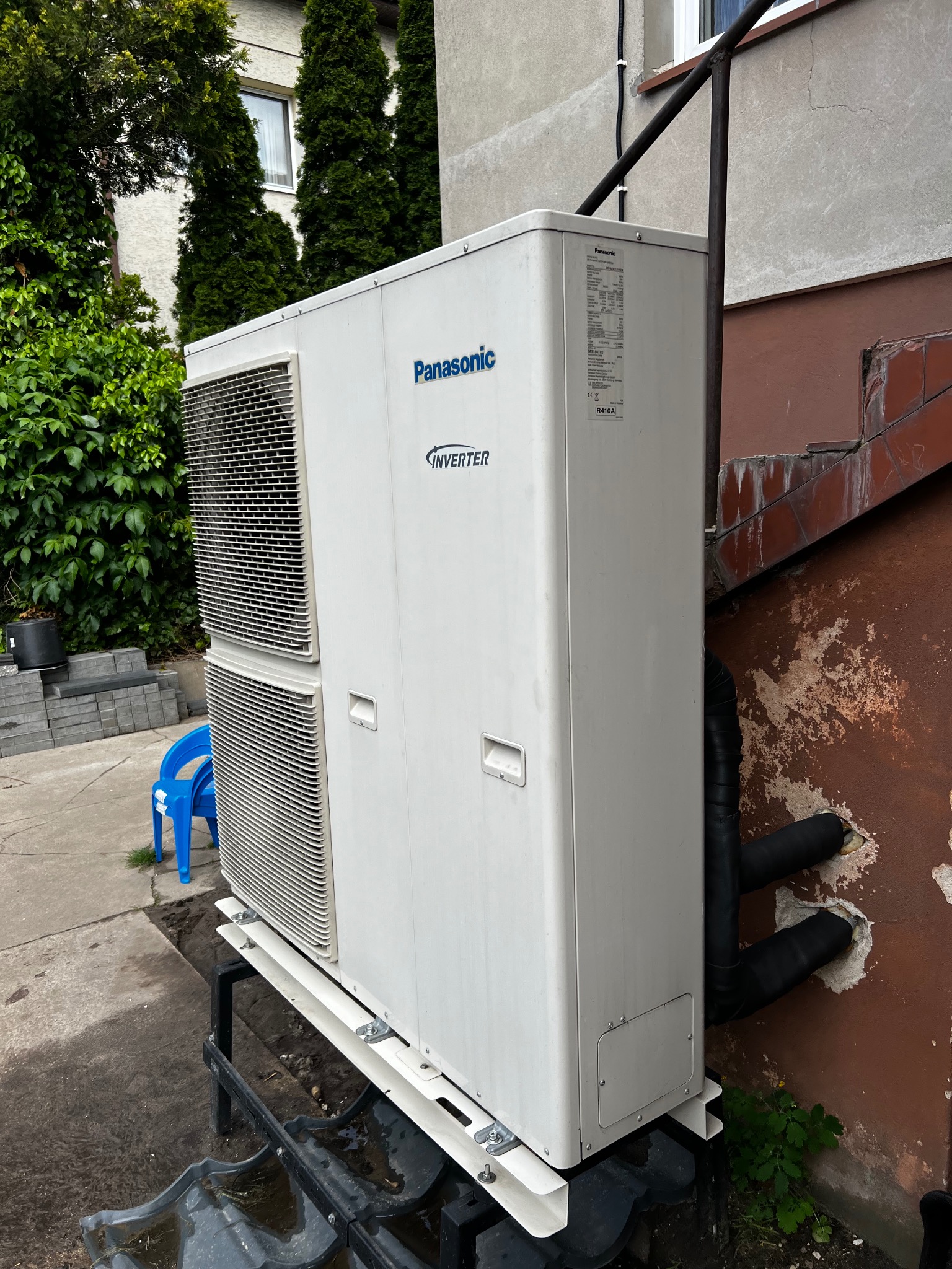 Zewnętrzna jednostka klimatyzacji Panasonic Inverter, zamontowana na metalowej konstrukcji przy ścianie budynku. Widoczne rury w izolacji wchodzące w ścianę.