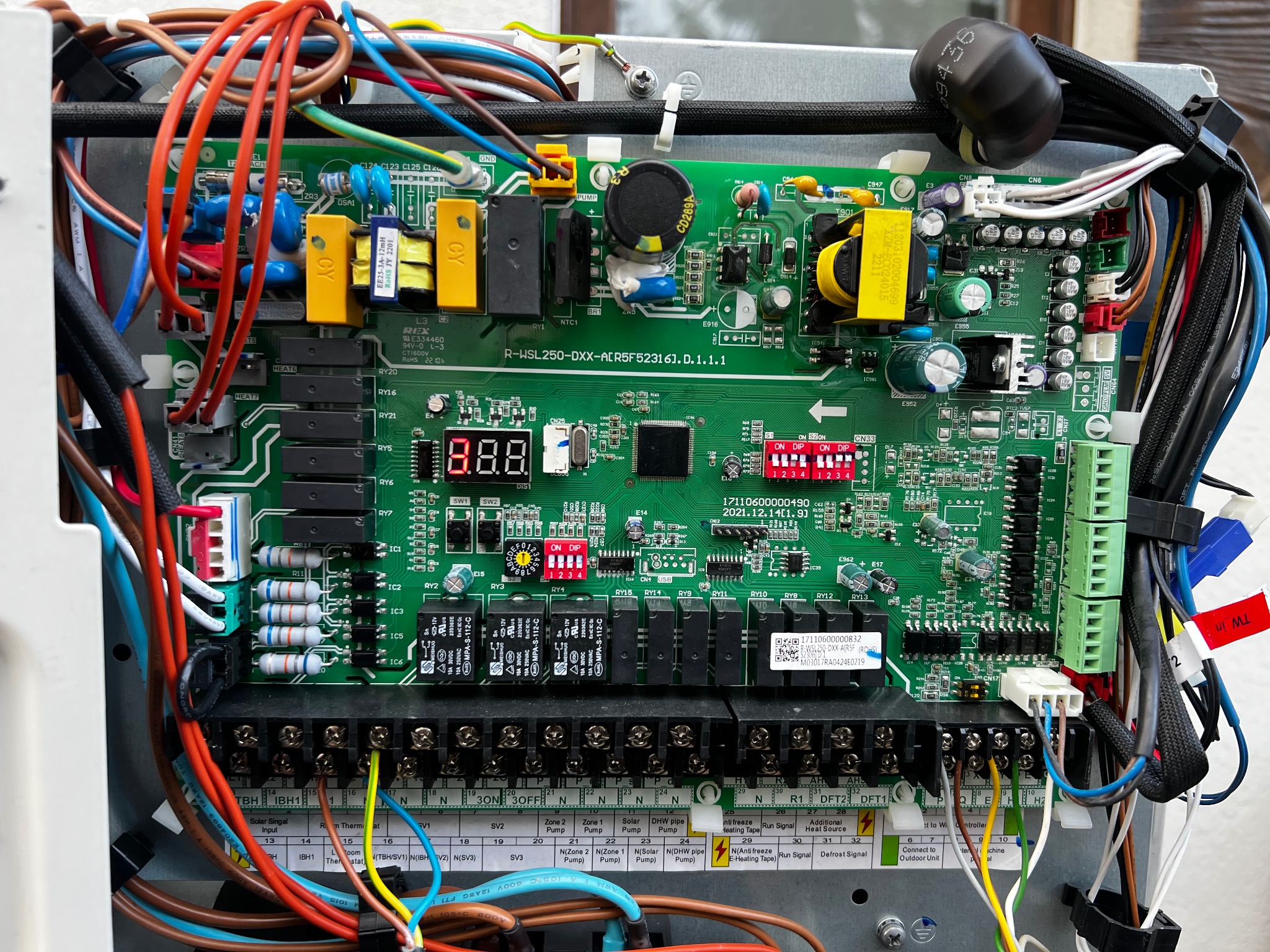 Szczegółowe ujęcie wewnętrznej elektroniki urządzenia, prawdopodobnie pompy ciepła, z widocznymi przewodami, płytką drukowaną i elementami sterującymi.