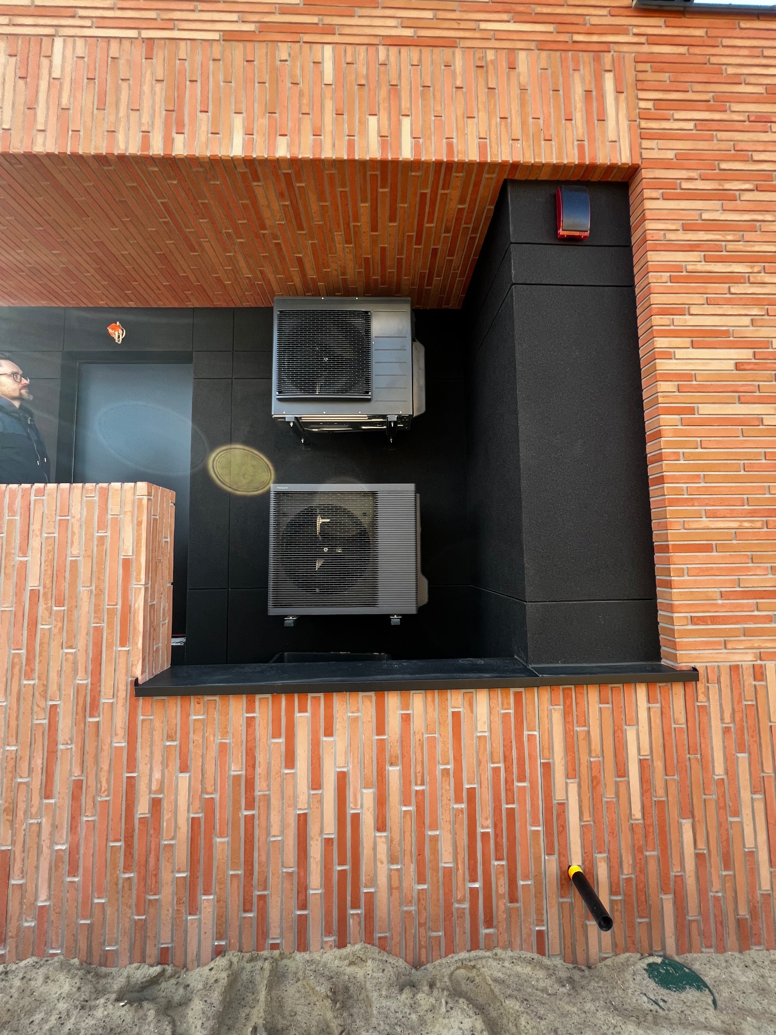 Dwa klimatyzatory zamontowane na elewacji z cegły klinkierowej, czarne panele pod urządzeniami. Widoczny fragment osoby w okularach po lewej stronie.