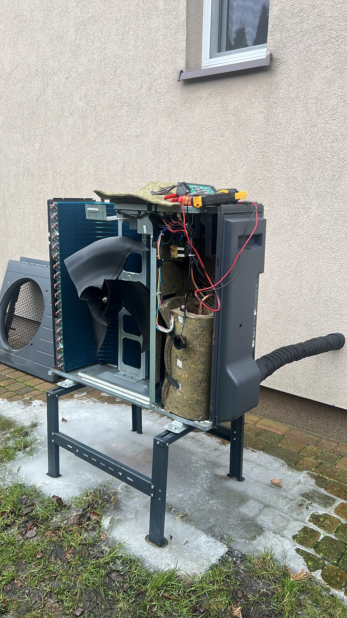 Otwarta obudowa pompy ciepła na zewnątrz budynku, widoczne elementy wewnętrzne i narzędzia diagnostyczne, otoczenie z resztkami śniegu.