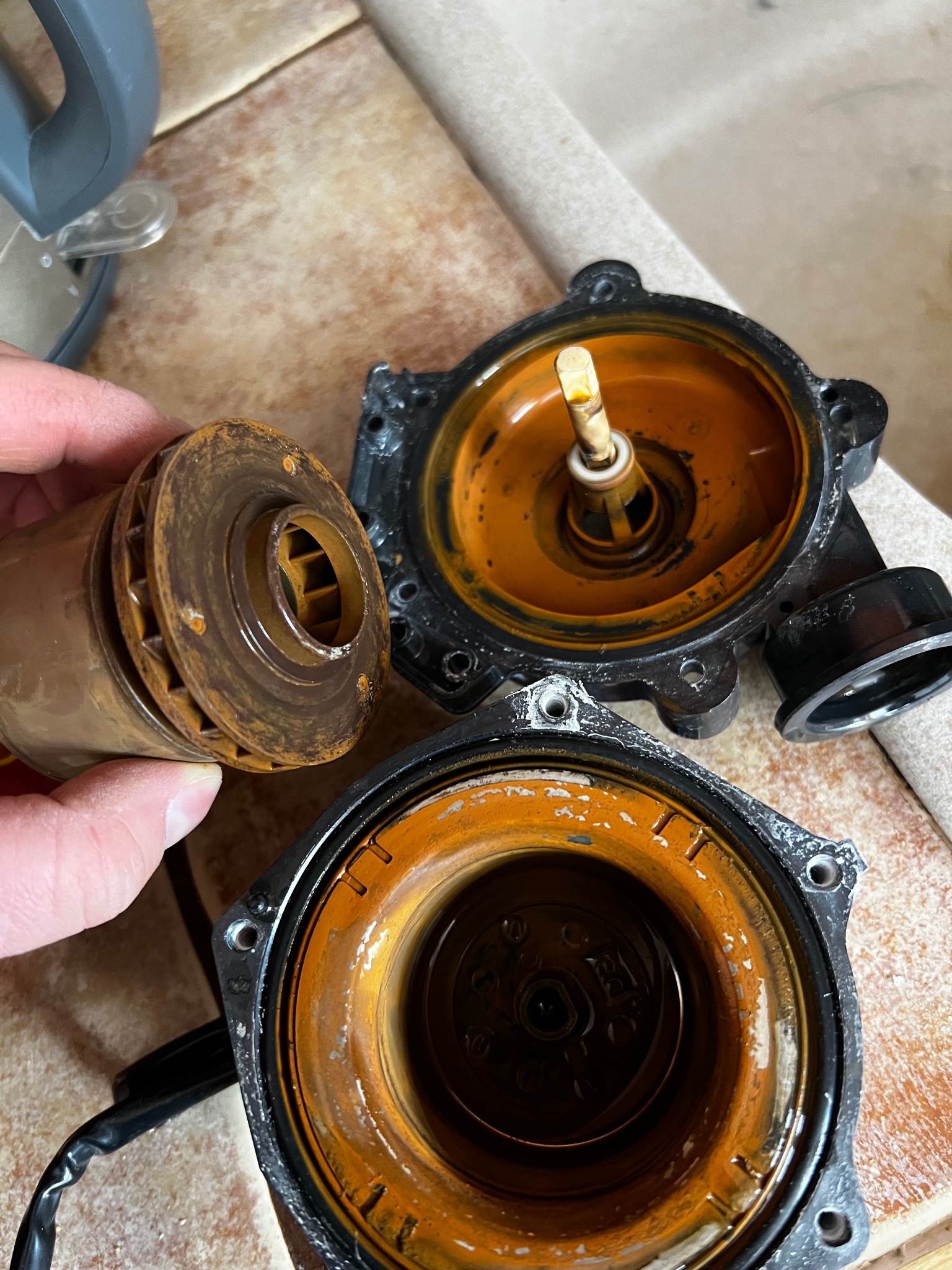 Zdemontowana, skorodowana pompa obiegowa z widocznym wirnikiem w dłoni, ukazująca zużycie i potrzebę serwisu lub wymiany. Elementy pompy na ceramicznej powierzchni.