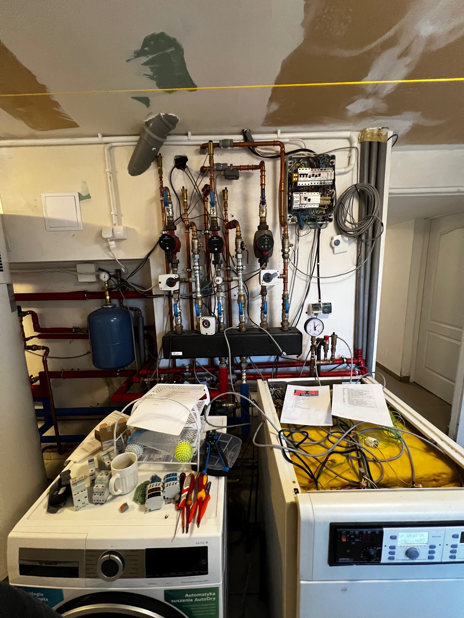 Węzeł cieplny z licznymi rurami miedzianymi, zaworami i manometrami, zamontowany na ścianie w pomieszczeniu technicznym. Widoczne elementy elektryczne i urządzenia.