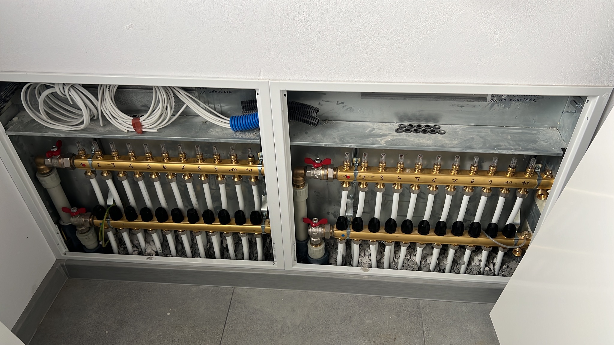 Rozdzielacz ogrzewania podłogowego w metalowej obudowie, widok z góry. Dwa rzędy rur z numeracją, przewody elektryczne w tle. Instalacja w kotłowni.