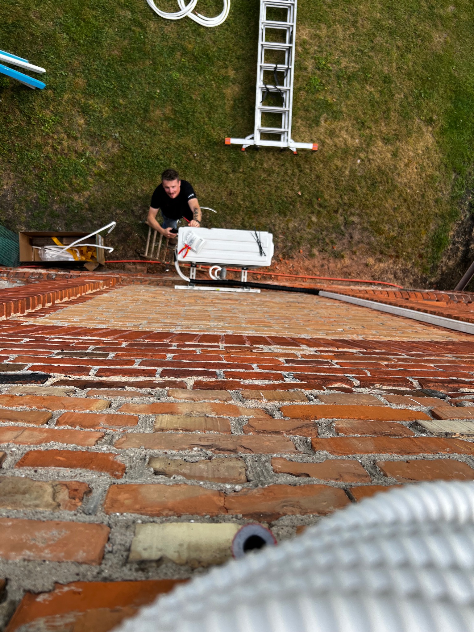 Montaż klimatyzacji na dachu z cegły. Instalator na drabinie przygotowuje jednostkę zewnętrzną. Widok z góry, z perspektywy dachu, na trawnik i narzędzia.