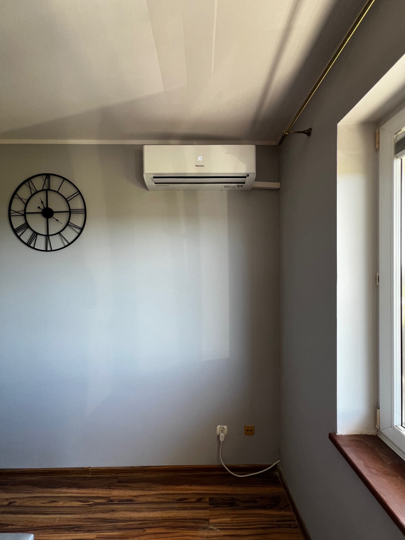 Biały klimatyzator ścienny w szarym pokoju z drewnianą podłogą, widoczny zegar na ścianie i okno z parapetem. Minimalistyczny design wnętrza.