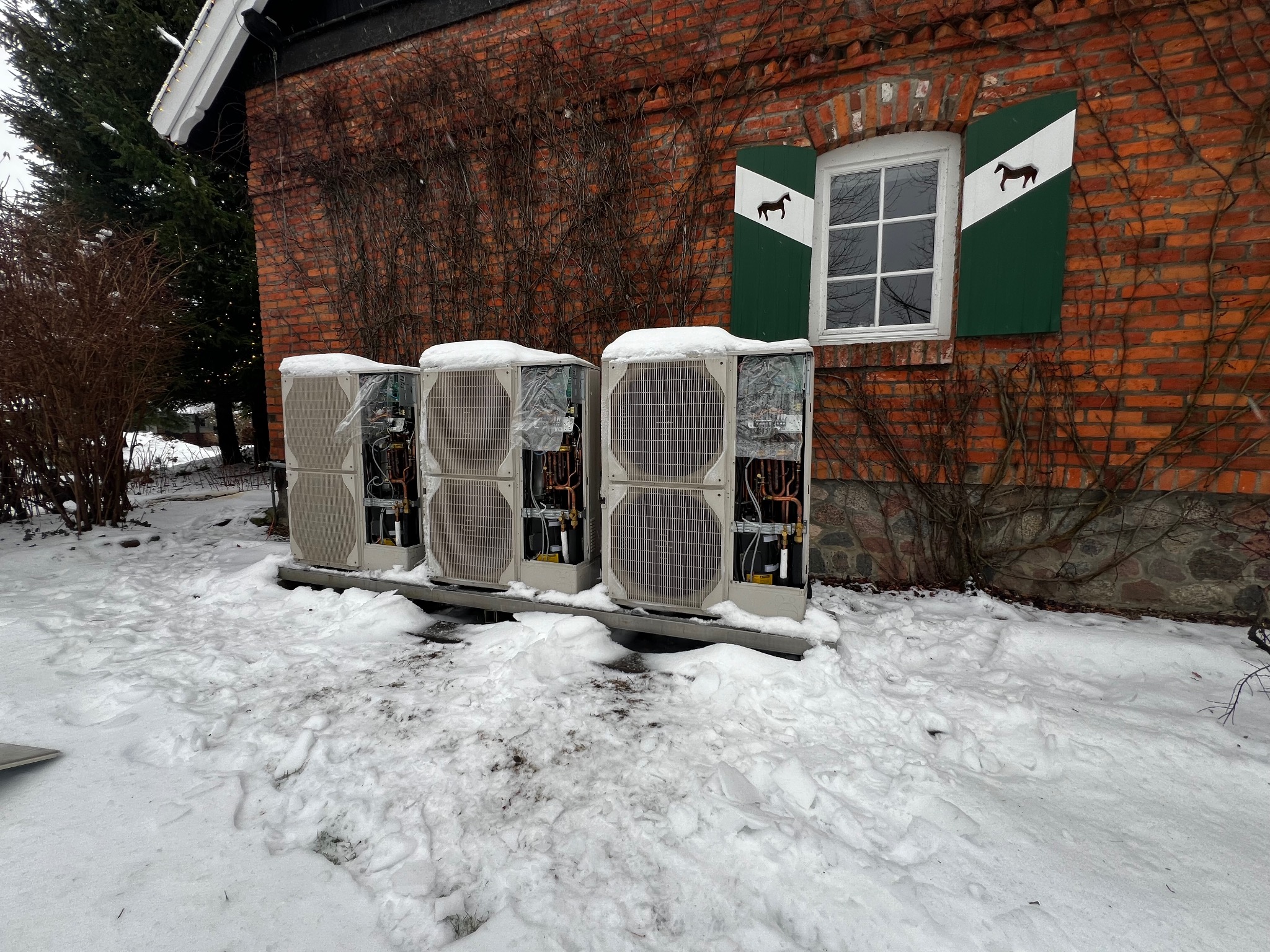 Trzy jednostki zewnętrzne pompy ciepła, częściowo zasypane śniegiem, ustawione przed ceglanym budynkiem z oknem z zielonymi okiennicami z motywem konia.