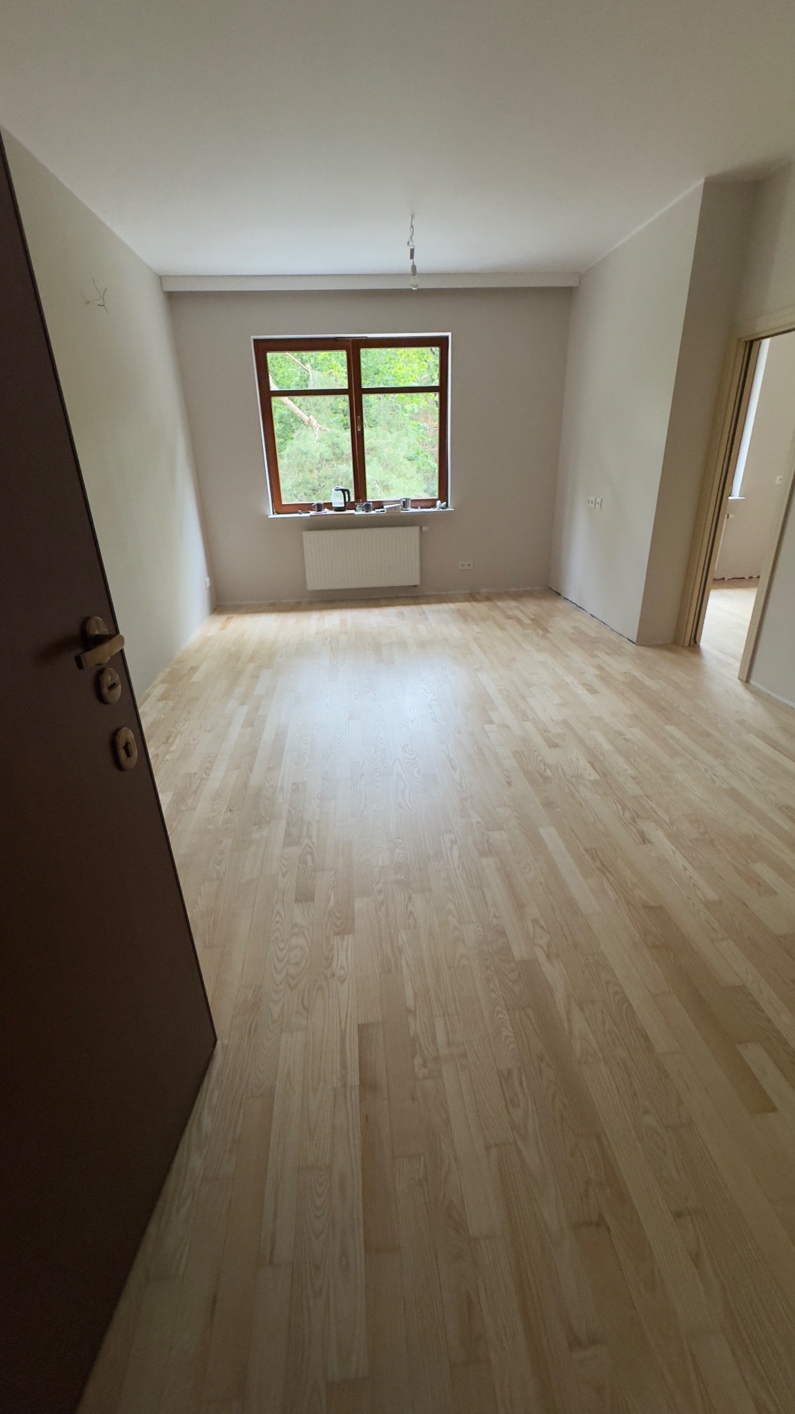 Jasny parkiet w pokoju z oknem i grzejnikiem. Widoczny fragment otwartych drzwi. Świeżo wykończona podłoga w neutralnej tonacji, gotowa do użytku.